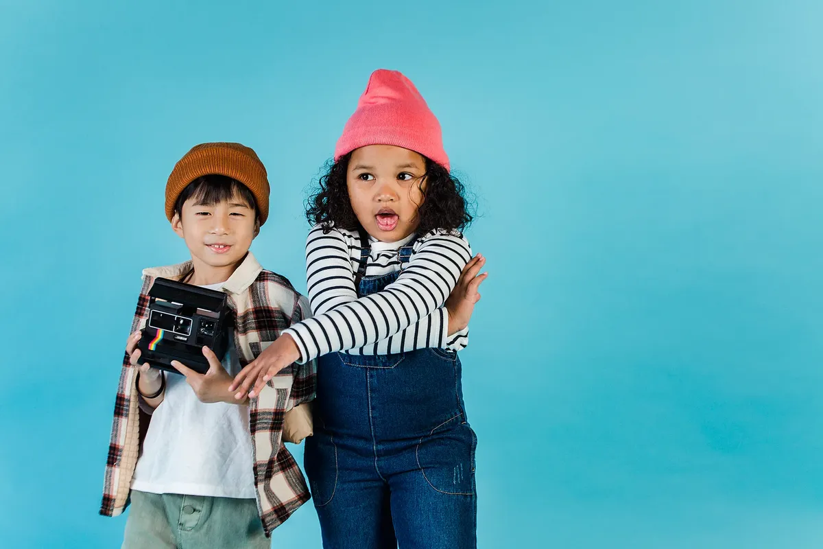 Cheerful little diverse children in trendy outfits standing with retro film photo camera against blue background in studio