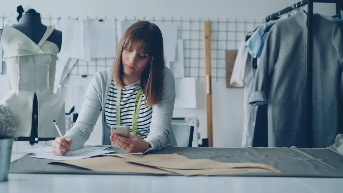 Fashion designer sketches patterns and uses smartphone in studio.